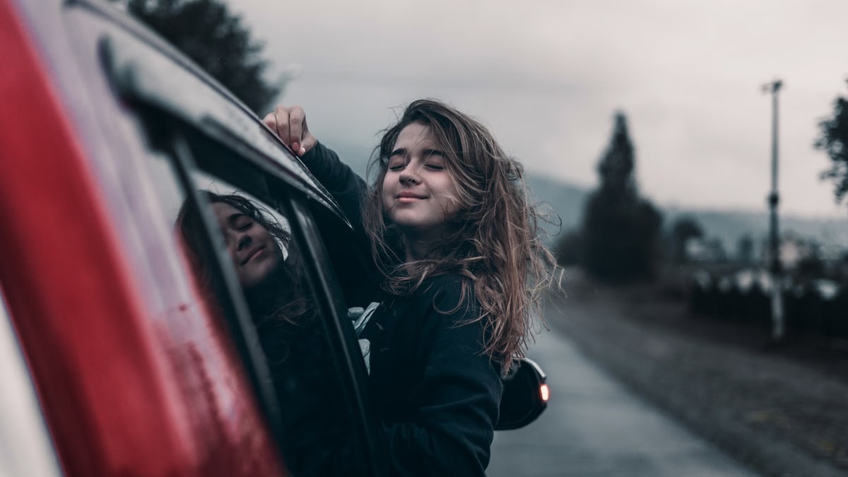 Side view of young dreamy ethnic female with closed eyes leaning out automobile window driving on asphalt road near trees and mountains under sky in countryside